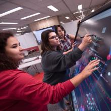 Three students work at a smart board in the Beta Classroom